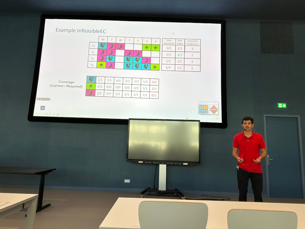 Roy presents in front of a big screen in a lecture room. The slide reads "Example InfeasibleEC" and shows a table that demonstrates an example NRP for one week, with four nurses and three types of shifts (morning, afternoon and night). It also shows constraints for each nurse in terms of maximum work load, maximum number of night shifts, and their preference w.r.t. how many sequential shifts they have. Below the table is an overview of the coverage constraints. The table itself contains a feasible solution.