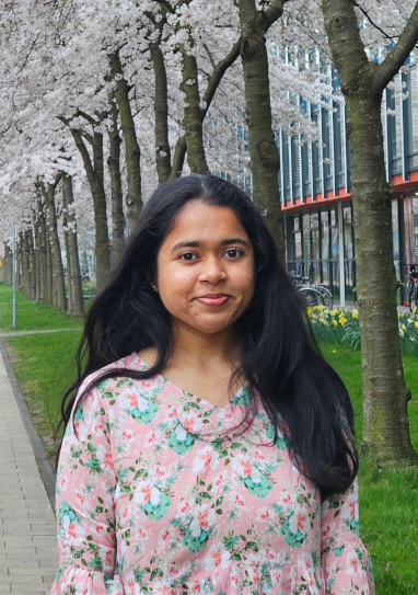 Professional photo of a young woman with black hair, wearing a pink-and-green flowery blouse. She's smiling at the camera, in front of a background of university buildings and blossoming trees.