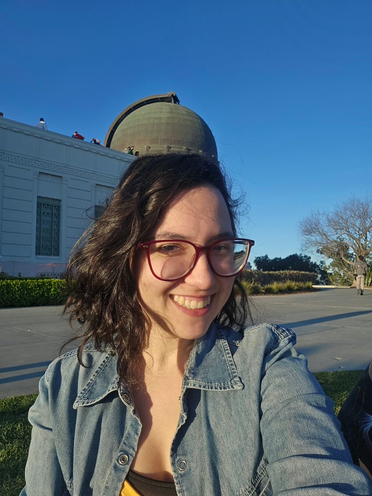 Casual photo of a young woman with very dark brown hair, wearing wine-red glasses and a denim jacket. She's smiling at the camera, in front of the cupola of an astronomical observatory against a clear blue sky.