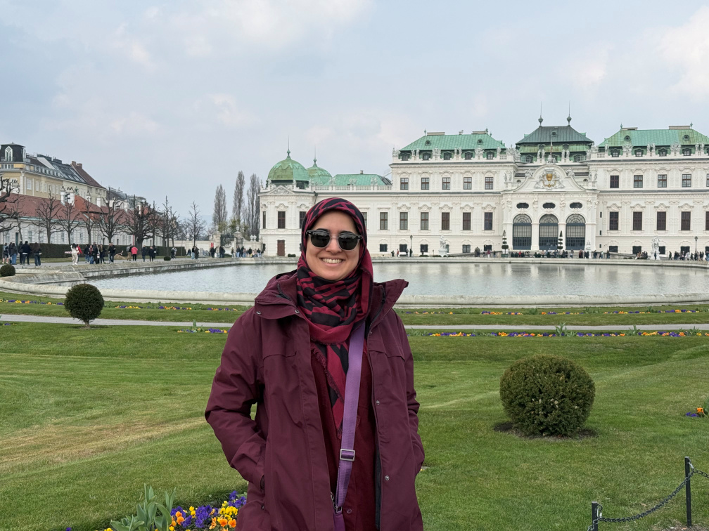 Casual photo of a young woman wearing sunglasses, a wine-red rain coat and a matching hijab. She's smiling at the camera, in front of a white palace with a copper-green roof, with a circular pond and carefully manicured lawn, decorated with colourful flowers.