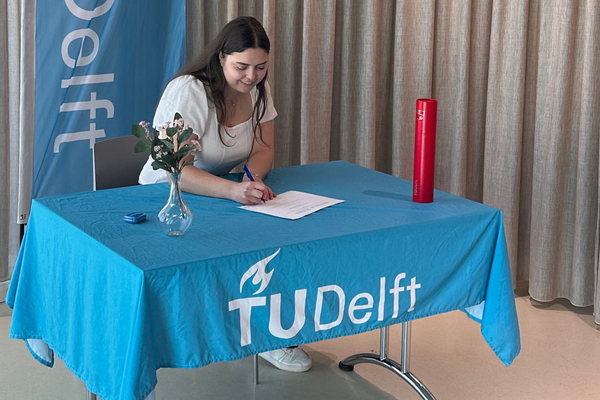 Roxana signs her diploma. She is smiling as she does it, and a red certificate tube that will hold the diploma is on the table next to her. The table is covered in a cyan blue TU Delft table cloth, and behind her is a cyan blue TU Delft banner.